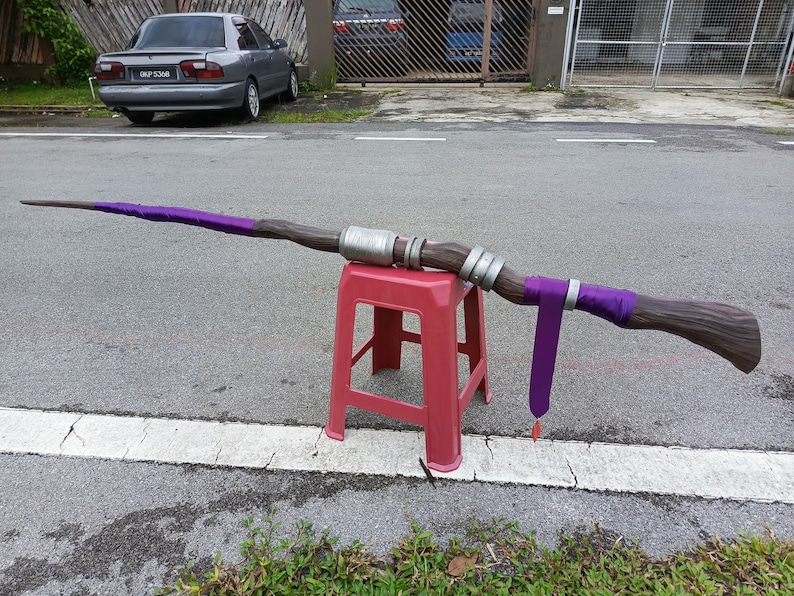 May include: A long, staff-like prop featuring a textured brown wood-grain finish, accented with metallic silver bands and vibrant purple fabric wraps. A long purple fabric strip hangs from the center, ending in a small, pointed red charm.