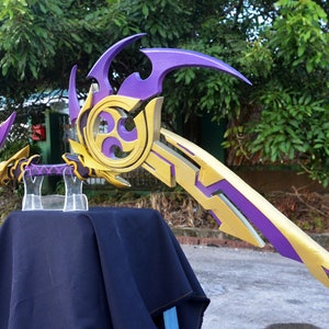 May include: A purple and gold bow and arrow prop with intricate details. The bow is curved and has a sharp point. The arrow is straight and has a feathered end. The prop is displayed on a black stand.