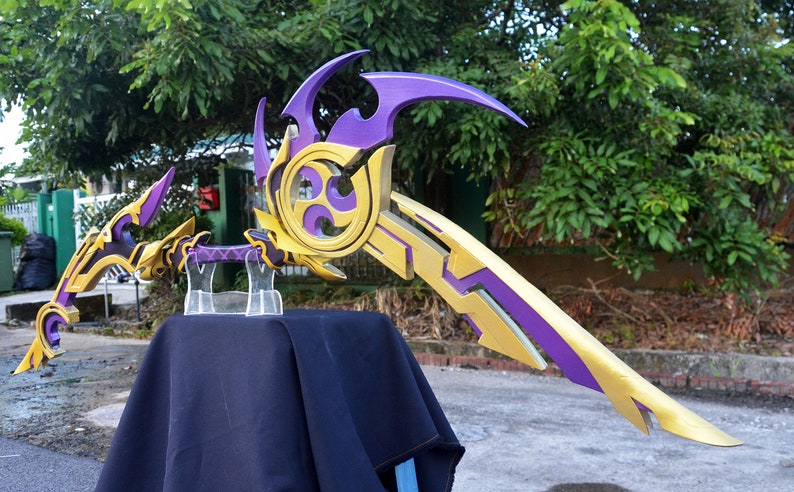 May include: A purple and gold bow and arrow prop with intricate details. The bow is curved and has a sharp point. The arrow is straight and has a feathered end. The prop is displayed on a black stand.
