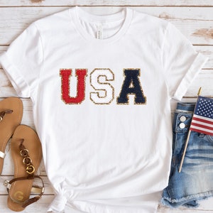 May include: White t-shirt with the word "USA" in red and navy blue letters with gold glitter trim. The shirt is paired with denim shorts, sandals, and a small American flag.