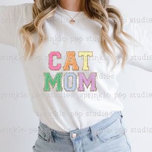 Puede incluir: Sudadera blanca con el texto "CAT MOM" en letras de colores brillantes y brillantes. Las letras est&aacute;n delineadas en oro.