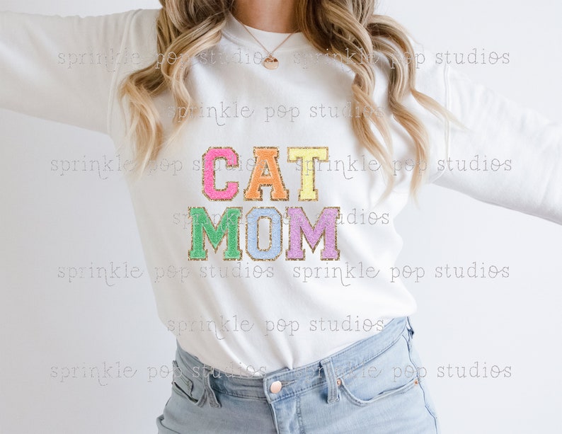 Puede incluir: Sudadera blanca con el texto "CAT MOM" en letras de colores brillantes y brillantes. Las letras est&aacute;n delineadas en oro.