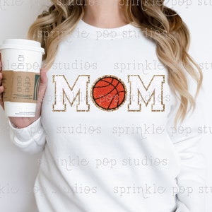Puede incluir: Sudadera blanca con letras de purpurina dorada que dicen "MOM" con una pelota de baloncesto en el medio de la palabra.