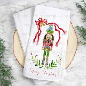 May include: A white kitchen towel featuring a watercolour illustration of a nutcracker holding a red ribbon with gold bells. The nutcracker is wearing a green and red suit with a blue hat. The towel has the text "Merry Christmas" printed on it.