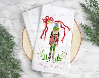 May include: A white kitchen towel with a watercolor illustration of a nutcracker holding a red ribbon with gold bells. The nutcracker is wearing a green and red suit with a blue hat. The towel has the text "Merry Christmas" printed on it.