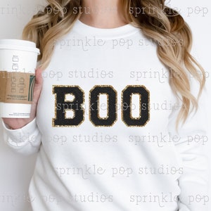 Może przedstawiać: Biała bluza z napisem "BOO" w czarnych literach z dodatkiem złotych brokatowych akcentów. Litery są w stylu uniwersyteckim.