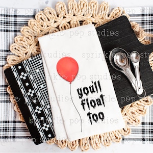 Könnte beinhalten: Weißes Küchentuch mit einem roten Ballon und dem Text "you'll float too" in Schwarz. Das Tuch liegt auf einer geflochtenen Unterlage mit einem schwarzen Schneidebrett und Messlöffeln.