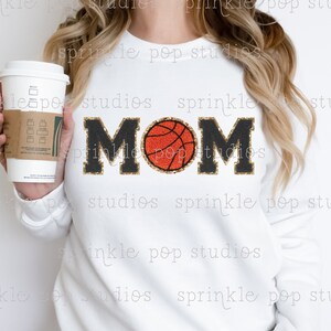May include: White sweatshirt with the word "MOM" in black letters outlined with gold glitter. A basketball replaces the "O" in "MOM". The sweatshirt is a casual style, perfect for a sports fan.