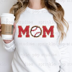 May include: White sweatshirt with the word "MOM" in red glitter letters with a gold glitter baseball in the centre of the word.