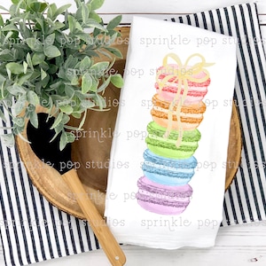 Op de afbeelding: Een witte keukendoek met een aquarelillustratie van een stapel kleurrijke macarons die met een geel lint zijn vastgebonden. De macarons zijn in regenboogkleuren gerangschikt, met de bovenste macaron roze, gevolgd door oranje, geel, groen, blauw en paars.