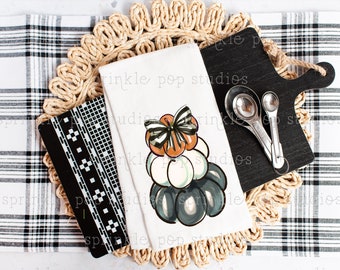 Puede incluir: Una toalla de cocina blanca con un fondo a cuadros blanco y negro. La toalla presenta una pila de calabazas con un lazo blanco y negro en la parte superior. Las calabazas están pintadas en tonos de naranja, blanco y gris. La toalla está sobre un mantel individual tejido con una tabla de cortar negra y cucharas medidoras.