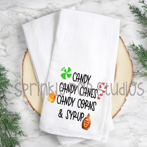 May include: White kitchen towel with a green candy cane, red candy cane, orange candy corn, and a bottle of maple syrup. The text reads "Candy Candy Canes Candy Corns & Syrup".