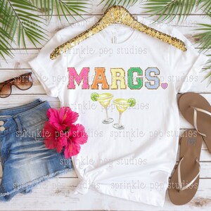 May include: White t-shirt with the text "MARGS." in rainbow glitter letters and two margarita glasses with lime slices.