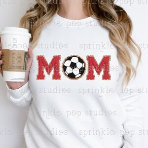 Puede incluir: Sudadera blanca con letras de purpurina roja que deletrean "MOM" y una pelota de fútbol de purpurina dorada en el centro.