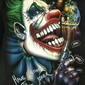 Puede incluir: Una camiseta negra con un gráfico de un payaso de pelo verde con una nariz roja y una gran sonrisa de dientes. El payaso sostiene una pistola y hay fuegos artificiales en el fondo. El texto "HAVE FUNTIME THEN JOKER" está impreso en la camiseta.