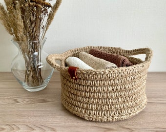 May include: A round, woven jute basket with two handles, holding rolled white, beige, and brown towels. A small leather tag is attached to the basket. A clear glass vase with dried plants is in the background.