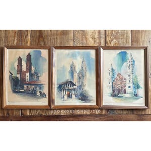 May include: Three framed watercolor paintings of European architecture. Each painting features a different building with a brown wooden frame. The paintings are mounted on a wooden board. The paintings have a vintage aesthetic.