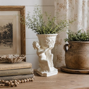 May include: A white ceramic vase featuring a sculpted cherub figure holding a fluted vessel, filled with green foliage. The vase sits near a stack of books, a small textured bowl, and a brown ceramic pot, with the text SOKYDESIGNSTUDIO visible.