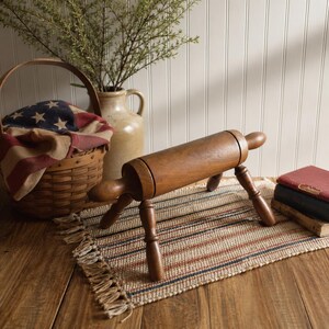 May include: A wooden rolling pin with a cylindrical body and rounded handles, supported by three angled legs. It rests on a striped rug with a fringed edge. A basket with an American flag is in the background.