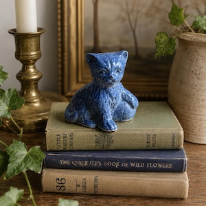May include: A small, blue, speckled ceramic cat figurine in a seated position. The cat has pointed ears, visible facial features, and a textured surface. It is placed on top of a stack of three vintage hardcover books with visible text.