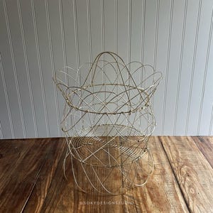 May include: A decorative, open-weave wire basket with a scalloped top and a handle. The basket is made of a light gold-colored metal and sits on a wooden surface. The basket's design is airy and open, suitable for storage or display.