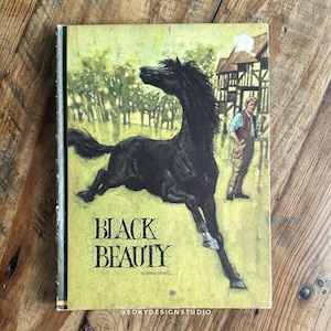Peut inclure: Un livre ancien intitul&eacute; "Black Beauty" par Anna Sewell. La couverture pr&eacute;sente un cheval noir galopant dans un champ, avec un homme et un b&acirc;timent en arri&egrave;re-plan. La couverture est jaune et verte avec du texte noir.