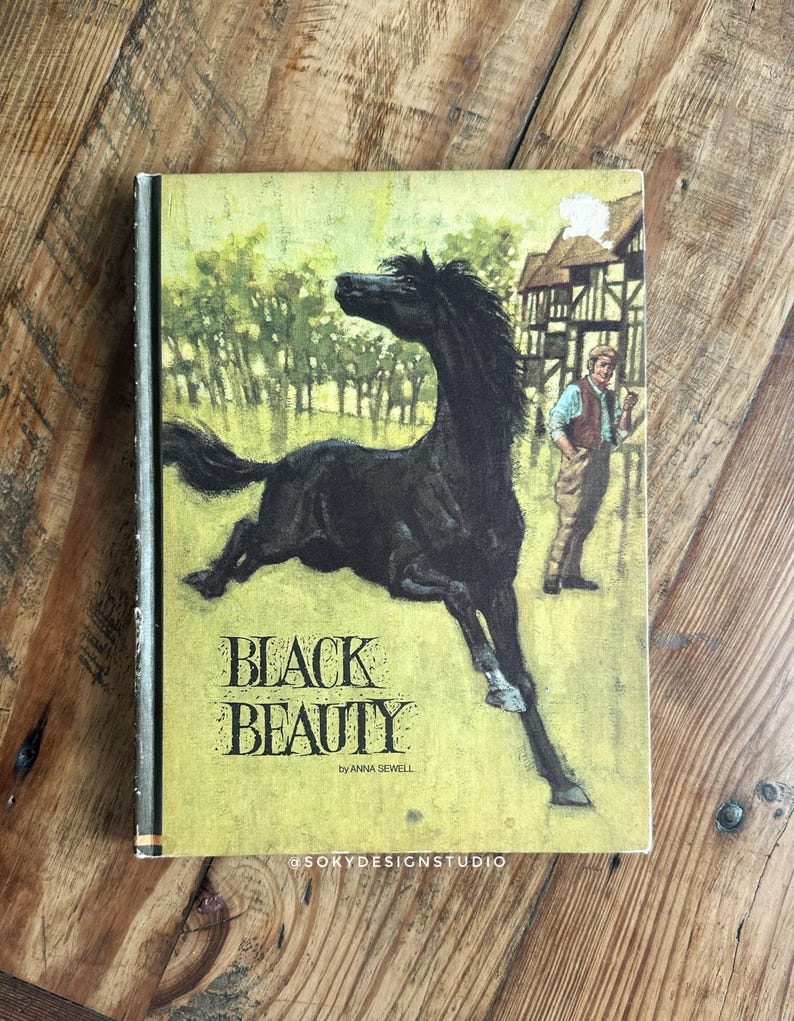 Peut inclure: Un livre ancien intitul&eacute; "Black Beauty" par Anna Sewell. La couverture pr&eacute;sente un cheval noir galopant dans un champ, avec un homme et un b&acirc;timent en arri&egrave;re-plan. La couverture est jaune et verte avec du texte noir.