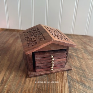 May include: A wooden house-shaped coaster holder with a carved floral design on the roof. The holder contains several square coasters. The coasters and holder are a dark brown color. The holder has a small brass knob.