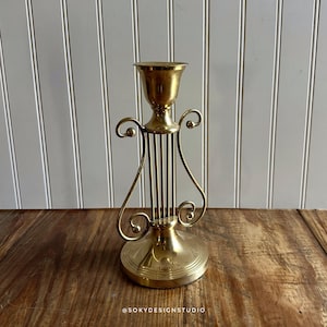 May include: A brass candlestick holder with a round base and a decorative, curved design. The holder features a cup-shaped top for holding a candle and vertical bars in the center. The candlestick is displayed on a wooden surface.