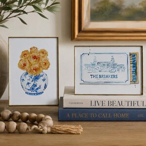 May include: Two framed art prints with gold edges. One print features a watercolor of yellow roses in a blue and white floral vase. The other print depicts a matchbox illustration labeled The Breakers Palm Beach with blue matches inside.