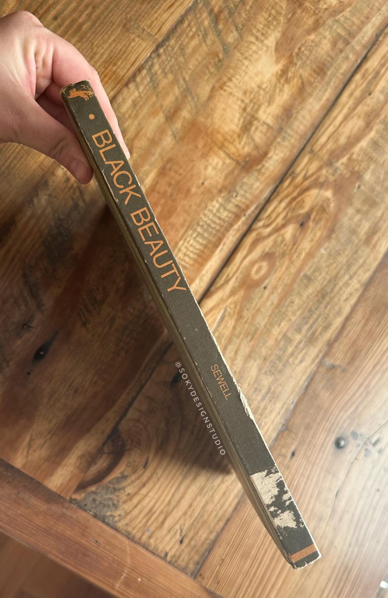 Peut inclure: Un exemplaire vintage du livre "Black Beauty" de Sewell. Le dos du livre est vert fonc&eacute; avec le titre en lettres orange. Le livre est pos&eacute; sur une surface en bois.