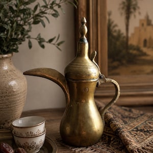 May include: A vintage brass coffee pot with a long spout and handle, next to two small white and gold cups on a brass plate. Dates and a decorative rug are also on the wooden table. A vase with greenery and a framed painting are in the background.
