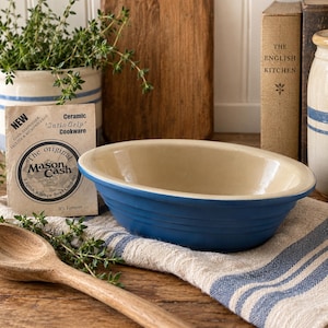 May include: An oval ceramic baking dish with a deep blue exterior and a smooth cream-colored interior. The dish features horizontal ribbed detailing along the sides. A paper tag labeled 'The original Mason Cash English Kitchen Bowl' is included.
