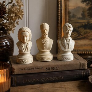 May include: Three off-white resin busts of composers standing on two stacked vintage books. The busts are labeled Bach, Tchaikovsky, and MacDowell. Each figure features detailed facial features and period-appropriate clothing, resting on a circular base.