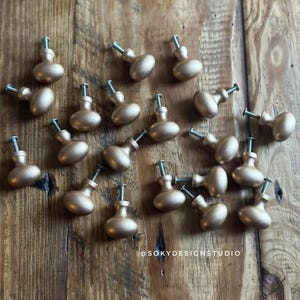 May include: A collection of gold-colored cabinet knobs with silver screws, arranged on a wooden surface. The knobs have an oval shape and a metallic finish. The image includes the text "@SOKYDESIGNSTUDIO".