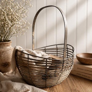 May include: A rustic metal wire basket with a curved handle, holding a folded beige cloth. The basket sits on a wooden surface, with a vase of white flowers and a wooden bowl in the background. The basket's design is simple and functional.