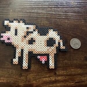 May include: A pixelated cow made of black, white, and tan beads. The cow has a pink udder and muzzle. A quarter is next to the cow for size comparison. The cow is on a dark brown wooden surface.