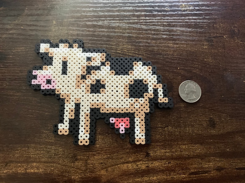 May include: A pixelated cow made of black, white, and tan beads. The cow has a pink udder and muzzle. A quarter is next to the cow for size comparison. The cow is on a dark brown wooden surface.