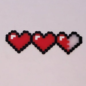 Puede incluir: Tres corazones pixelados, dos son rojos y uno es mitad rojo y mitad blanco. Los corazones est&aacute;n hechos de peque&ntilde;as cuentas.