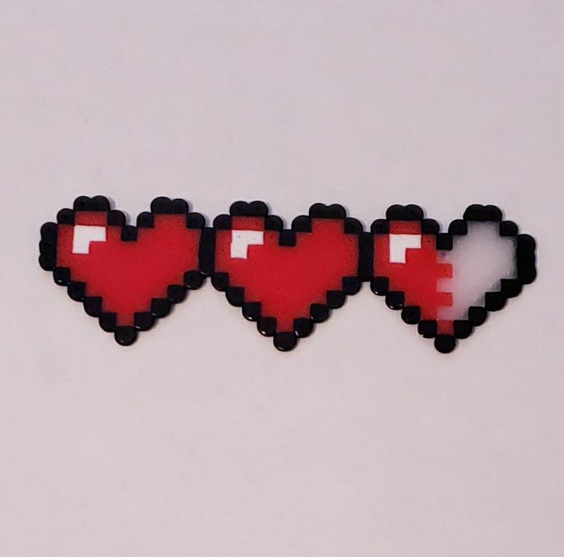 Puede incluir: Tres corazones pixelados, dos son rojos y uno es mitad rojo y mitad blanco. Los corazones est&aacute;n hechos de peque&ntilde;as cuentas.