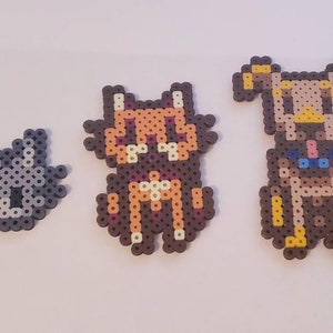 May include: Three handmade bead creations: a gray cat, a brown and orange cat, and a brown and tan dog. Each animal is made with small, colorful beads, and they are arranged in a row. The cat and dog figures are sitting.