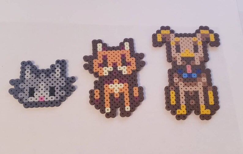 May include: Three handmade bead creations: a gray cat, a brown and orange cat, and a brown and tan dog. Each animal is made with small, colorful beads, and they are arranged in a row. The cat and dog figures are sitting.