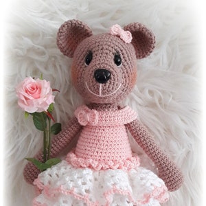 Op de afbeelding: Een gehaakte bruine beer die een roze jurk draagt met witte kant en een roze roos vasthoudt. De beer heeft bruine ogen en een zwarte neus. De beer draagt witte sokken en roze schoenen.