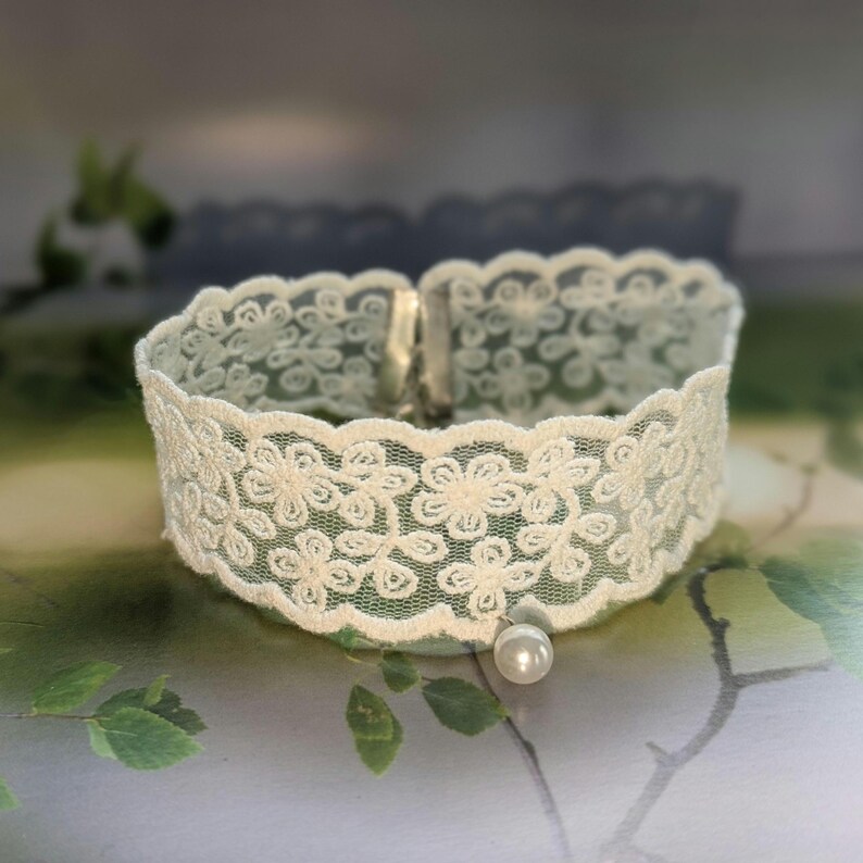 Pu&ograve; includere: Girocollo in pizzo avorio con motivo floreale e un piccolo pendente di perla. Il delicato pizzo &egrave; assicurato con una chiusura. Il girocollo &egrave; largo circa 2,5 cm.