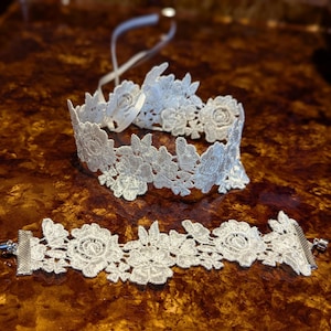 May include: A two-piece white lace floral jewellery set. The set includes a choker necklace with a ribbon tie and a bracelet with a metal clasp. The lace features a detailed floral pattern, creating an elegant and delicate appearance.