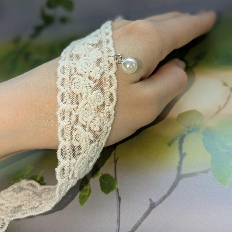 Pu&ograve; includere: Bracciale in pizzo avorio con motivo floreale e un ciondolo a perla pendente. Il delicato pizzo &egrave; avvolto attorno al polso, mettendo in mostra il design. La perla aggiunge un tocco di eleganza a questo accessorio.