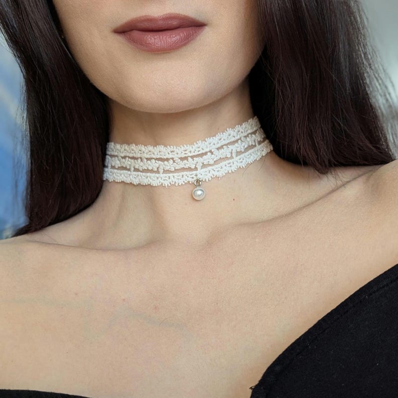 Pu&ograve; includere: Una delicata collana girocollo in pizzo bianco con un design a strati. La collana presenta un piccolo pendente di perla pendente. Il girocollo &egrave; indossato intorno al collo, mostrando il suo intricato motivo floreale e il suo stile elegante.