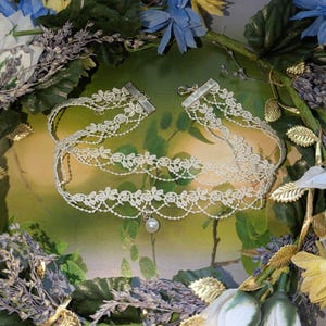 Pu&ograve; includere: Una delicata collana girocollo in pizzo bianco sporco con un pendente a perla. La collana presenta pi&ugrave; strati di pizzo floreale e chiusure argentate. La collana &egrave; esposta su uno sfondo di verde e fiori.