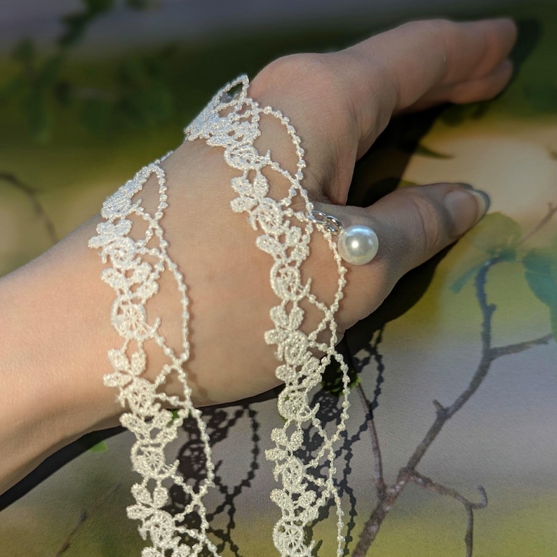 Pu&ograve; includere: Delicato braccialetto in pizzo bianco con motivi floreali e un grande accento di perla rotonda. Il braccialetto &egrave; collegato a un anello con una piccola catena d'argento. Il design &egrave; intricato ed elegante, adatto alle occasioni speciali.