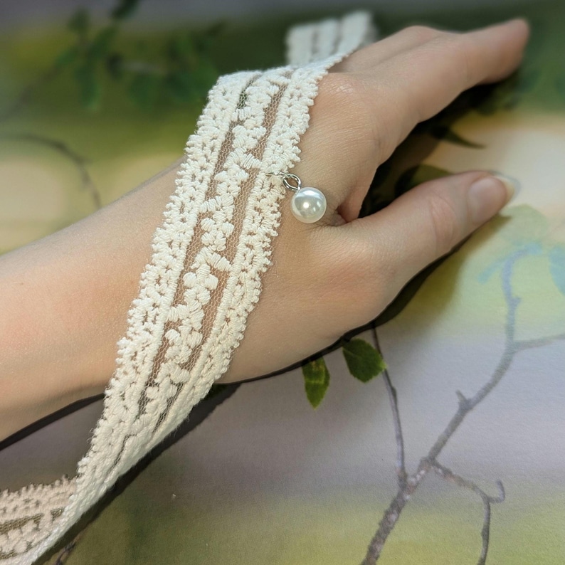 Pu&ograve; includere: Bracciale in pizzo color avorio con un delicato motivo floreale. Un piccolo ciondolo a perla pende. Il bracciale &egrave; progettato per essere indossato intorno al polso, offrendo uno stile vintage o boh&eacute;mien.